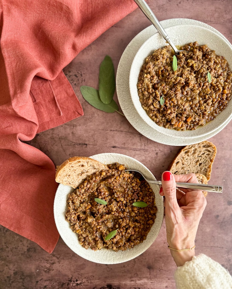 2 white bowls of Italian Lentil soup on a brown table with a hand collecting a spoon of the soup.