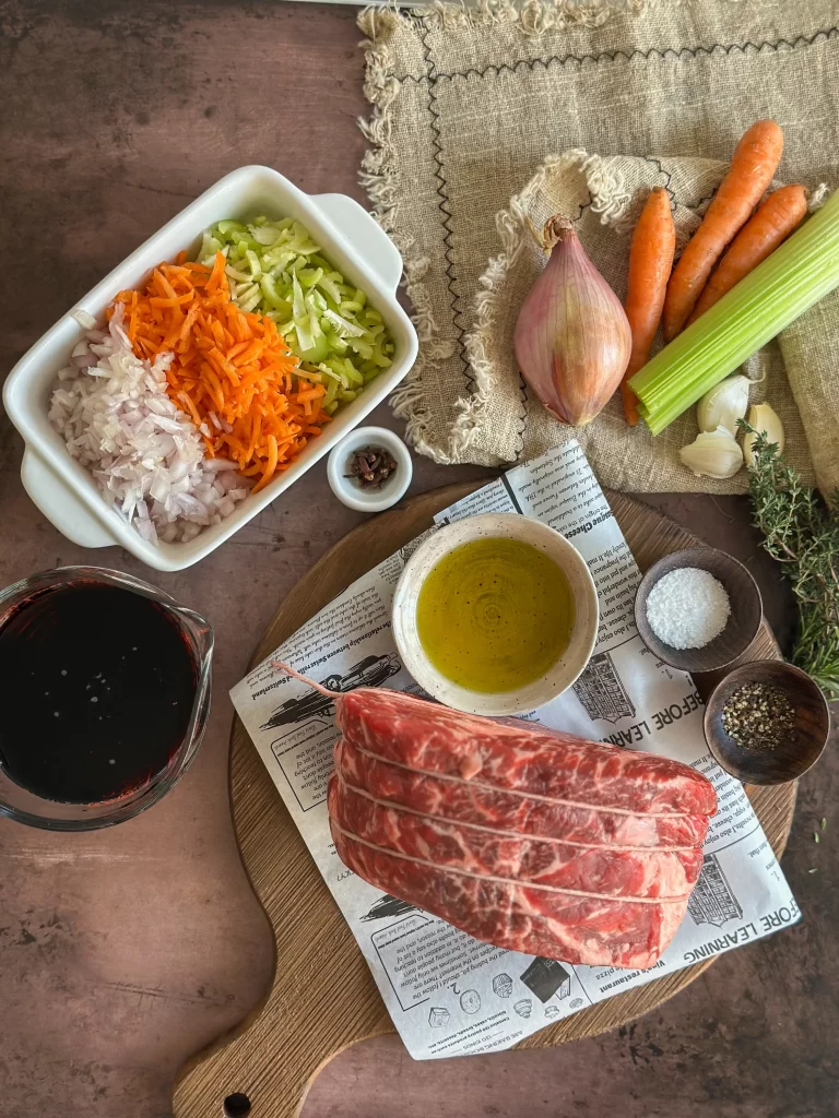 Ingredients for Brasato al Barolo on a table: carrots, celery and onions diced, garlic cloves, cloves, herbs, olive oil, salt and pepper, beef chuck, Barolo wine.