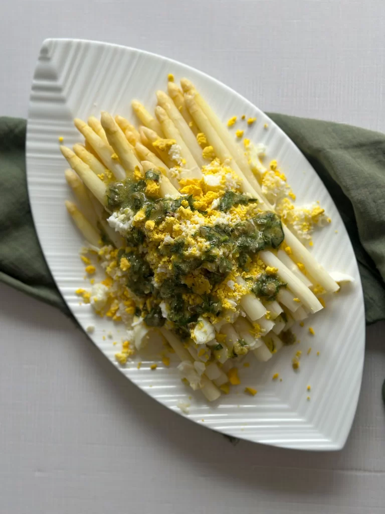 White asparagus topped with eggs ad capers vinaigrette on a white oval plate over a green napkin
