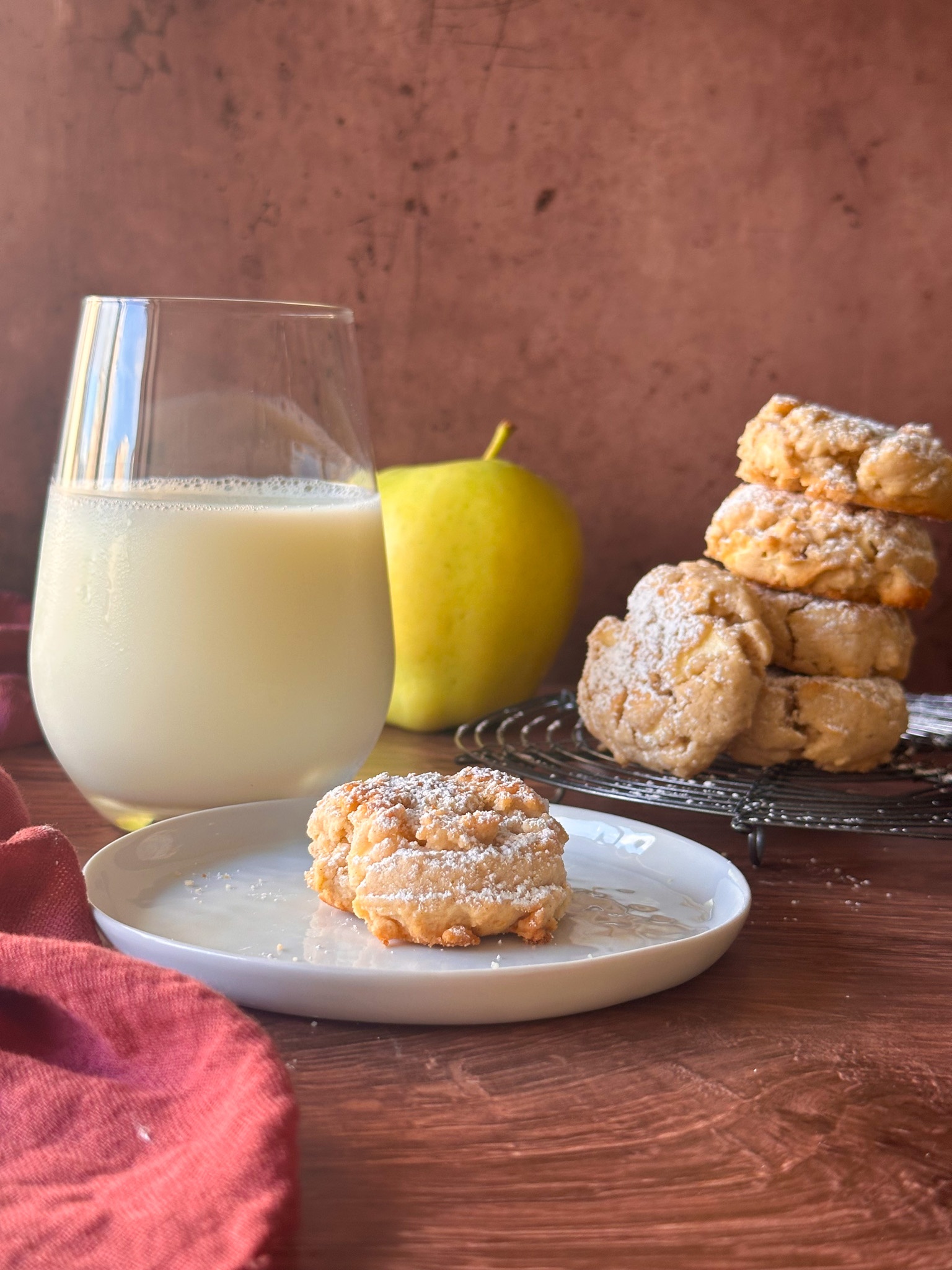 Cookie on a small round plate, with a cookie tower in the back alng with an apple, and a glass of milk